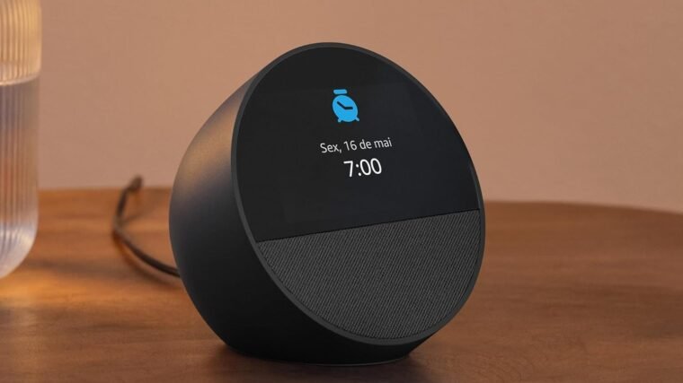 Imagem com o dispositivo Echo Spot sobre uma mesa. O dispositivo é um semi-círculo, com metade da face frontal dedicada a um painel que mostra horário e outras informações, e a metade de baixo sendo um alto-falante.