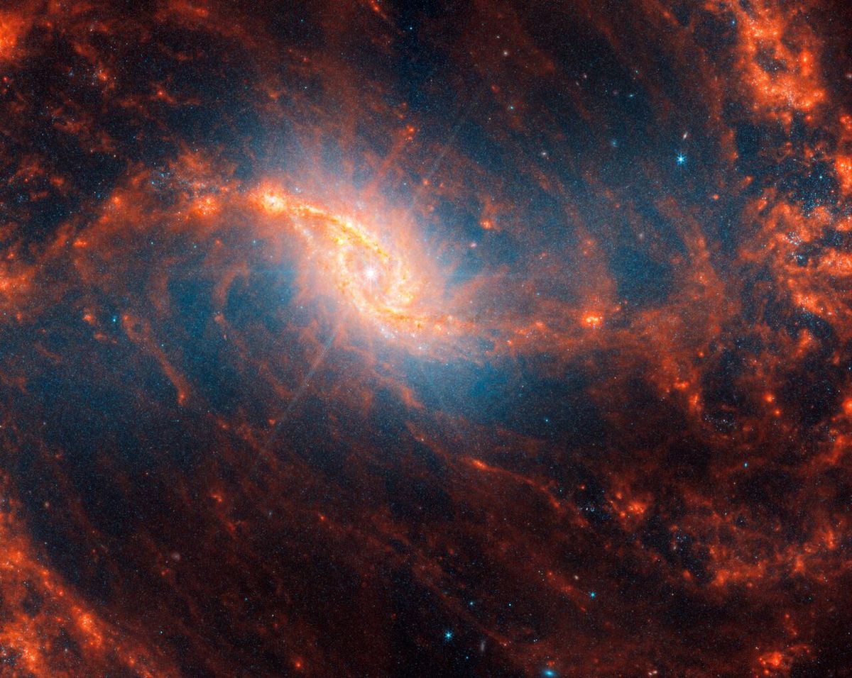This spiral galaxy was observed as part of the Physics at High Angular resolution in Nearby GalaxieS (PHANGS) program, a large project that includes observations from several space- and ground-based telescopes of many galaxies to help researchers study all phases of the star formation cycle, from the formation of stars within dusty gas clouds to the energy released in the process that creates the intricate structures revealed by Webb’s new images. NGC 1365 is 56 million light-years away in the constellation Fornax. Learn more about what can be seen in this vast collection of Webb images here. [Image description: Webb’s image of galaxy NGC 1365 shows a central region that looks like an angled, smashed oval that is a mix of bright orange shades that emanate a blue glow. The galaxy’s bar extends horizontally from the oval. It is crossed by messy dust filaments that curve slightly, forming a backward S shape. Fainter filaments appear below and above the core.]