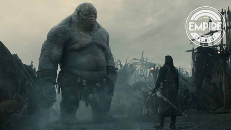 Foto da 2ª temporada de O Senhor dos Anéis: Os Anéis de Poder, com o Troll Damrod em destaque. O personagem fica de frente com um guerreiro em uma vila destruída pelo fogo.